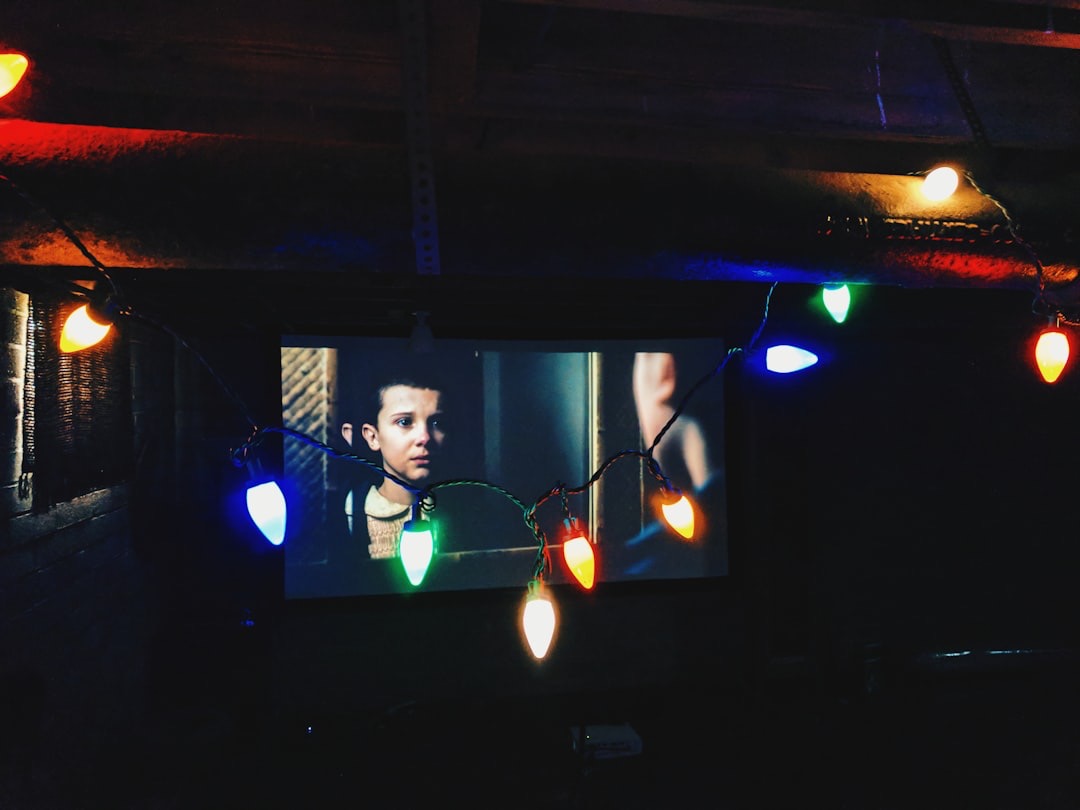 String lights hang before a TV playing a Stranger Things scene of Eleven staring at her mirror reflection